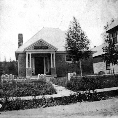 Black and white photo of a small, single-story library building with columns at the entrance, surrounded by a grassy yard and stone markers.