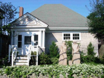 A quaint public library with shingled siding and a gabled roof. It features white columns at the entrance, surrounded by green shrubs and blooming flowers, creating a welcoming and serene atmosphere.