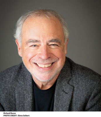 An older man with short gray hair and a beard smiles at the camera, wearing a gray blazer and black shirt against a plain background.